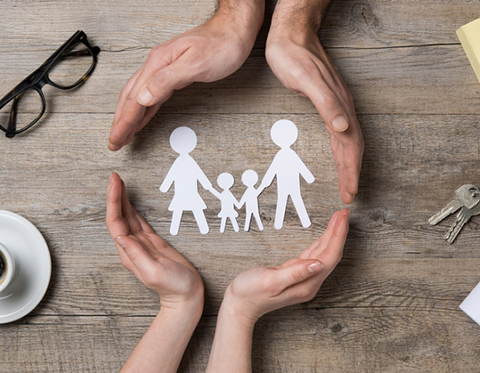 Two pairs of hands creating a circle around a paper cut out of a family. This illustrates that the family is protected.
