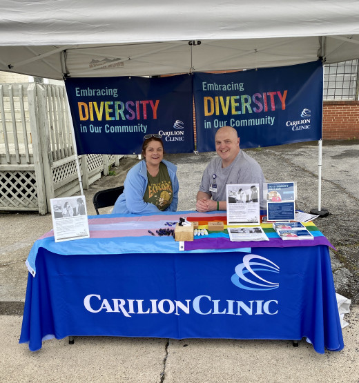 Photo of two members of Carilion's LGBTQ+ ERG sitting at a table with Carilion banners, information and giveaways at Roanoke's 30th annual PRIDE Festival
