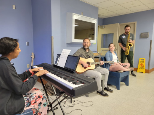 Karina Espinal, seated playing the keyboard, Eric Stachelski, seated playing the guitar, Laura Cuadrado seated singing, and Clay Trinkle standing playing the saxophone. 